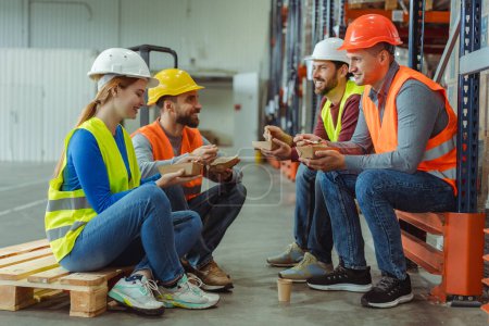 Photo for Group of happy workers, managers wearing hard hats, vests, holding lunch boxes, eating lunch, sitting, talking to each other during break. Concept of lunch together - Royalty Free Image
