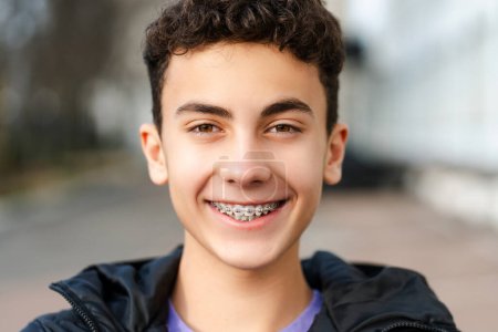 Téléchargez les photos : Portrait rapproché d'un beau garçon adolescent souriant avec des bretelles portant des vêtements décontractés, regardant la caméra, debout dans la rue urbaine. Concept dentaire, traitement, soins de santé - en image libre de droit