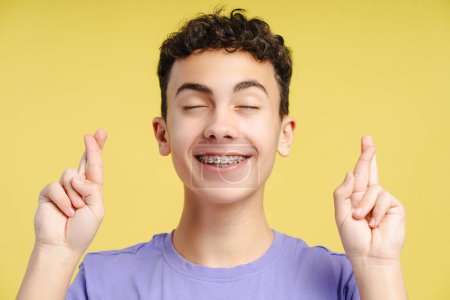 Téléchargez les photos : Adolescent attrayant souriant avec bretelles avec les yeux fermés et les doigts croisés espérant, rêvant, souhaitant, debout isolé sur fond jaune. Concept de faire un voeu - en image libre de droit