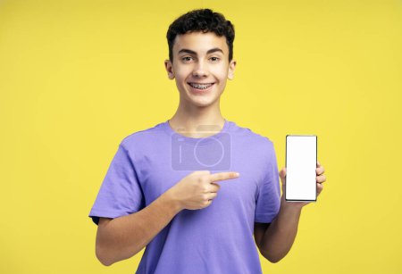 Foto de Guapo niño sonriente, adolescente de 15 años con aparatos ortopédicos, sosteniendo el teléfono móvil, utilizando la aplicación móvil, señalando con el dedo a la pantalla blanca en blanco, maqueta, espacio de copia. Concepto de publicidad, tecnología en línea - Imagen libre de derechos