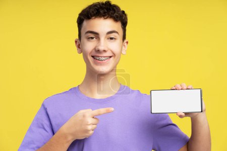 Photo for Attractive happy boy, teenager with braces, holding mobile phone, using mobile app, pointing finger at white blank screen, mockup, copy space isolated on yellow background - Royalty Free Image