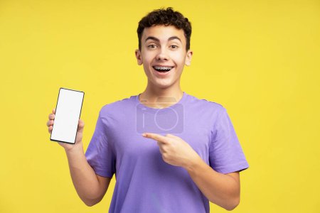 Foto de Sonriente, niño emocionado, adolescente sosteniendo el teléfono móvil, señalando con el dedo en blanco, pantalla blanca mirando a la cámara de pie aislado en el fondo amarillo. Concepto de tecnologías en línea, publicidad - Imagen libre de derechos