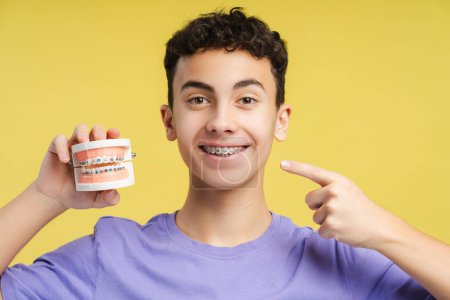 Photo for Handsome, smiling boy, teenager with braces holding jaw and pointing finger behind his teeth looking at camera while standing on yellow background. Concept of dental care, health care - Royalty Free Image