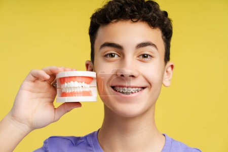 Photo for Confident, attractive boy, teenager with braces holding orthodontic jaw, looking at camera standing isolated on yellow background, closeup. Dental, treatment concept - Royalty Free Image