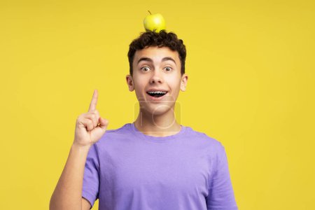 Téléchargez les photos : Garçon excité souriant, adolescent tenant la pomme sur la tête, doigt en l'air, ayant l'idée de regarder la caméra debout isolé sur fond jaune. Concept d'éducation - en image libre de droit
