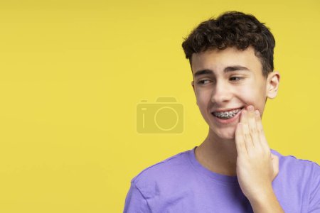 Téléchargez les photos : Garçon énervé, adolescent avec des bretelles touchant la joue ayant mal aux dents, regardant loin debout isolé sur fond jaune. Concept de soins dentaires, publicité, traitement - en image libre de droit
