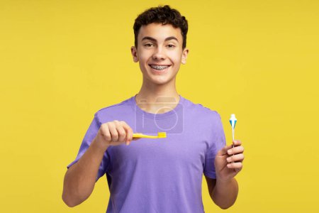 Foto de Muchacho atractivo y sonriente con frenos que sostienen el cepillo de dientes y la pasta de dientes mirando a la cámara. El adolescente positivo se cepilla los dientes. Concepto dental rutina matutina - Imagen libre de derechos
