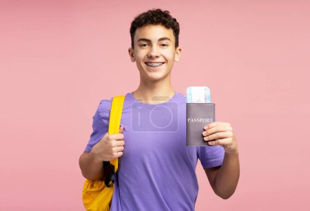 Smiling handsome boy, teenager holding passport and boarding pass, backpack looking at camera isolated on pink background. Concept of travel, excursion