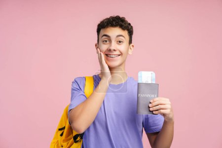 Excited boy, teenager holding passport and boarding pass, backpack looking at camera isolated on pink background. Concept of travel, excursion