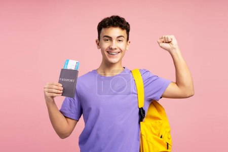 Attraktiver, aufgeregter Junge, Teenager mit Pass und Bordkarte, Rucksack auf der Schulter, glücklich, schaut vereinzelt in die Kamera auf gelbem Hintergrund. Konzept von Reisen, Urlaub