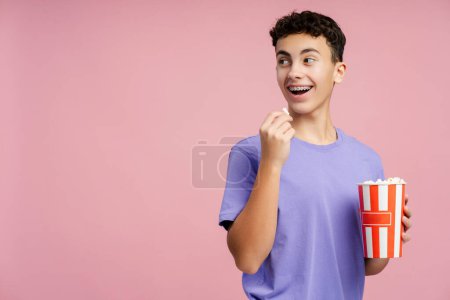 Foto de Sonriente adolescente atractivo comiendo palomitas de maíz, sosteniendo cubo mirando hacia otro lado en el espacio de copia de pie aislado sobre fondo rosa. Un chico guapo con aparatos ortopédicos viendo películas. Concepto de entretenimiento - Imagen libre de derechos