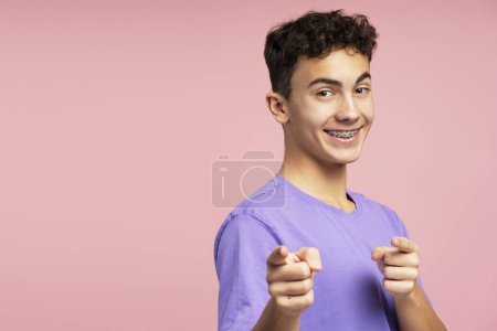 Téléchargez les photos : Adolescent attrayant beau avec des bretelles portant élégant tenue décontractée pointant des doigts, copier l'espace, regardant la caméra debout isolé sur fond rose. Concept de publicité - en image libre de droit
