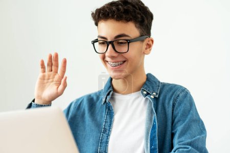 Téléchargez les photos : Adolescent garçon heureux souriant, étudiant avec des accolades portant des lunettes élégantes et des vêtements décontractés agitant la main, ayant un appel vidéo assis à table, utilisant un ordinateur portable. Concept d'éducation en ligne - en image libre de droit
