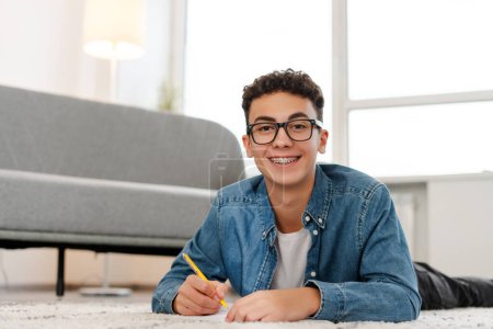 Photo for Attractive, happy boy, teenager wearing stylish eyeglasses lying on floor in room at home, taking notes, doing homework, looking at camera. Education concept - Royalty Free Image