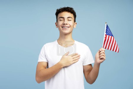 Téléchargez les photos : Adolescent patriote souriant attrayant jeune garçon tenant drapeau américain avec les yeux fermés tenant la main sur le cœur debout isolé sur fond bleu. Concept du 4 juillet, Jour de l'indépendance - en image libre de droit
