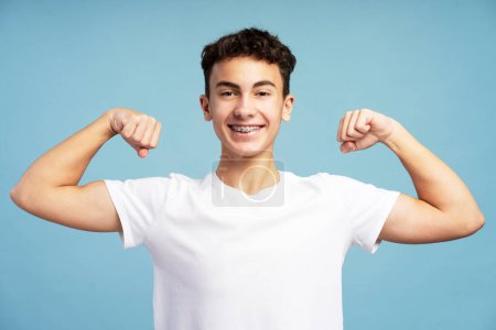 Photo for Smiling strong teenage boy wearing stylish white t shirt showing muscles looking at camera isolated on blue background. Advertisement concept - Royalty Free Image