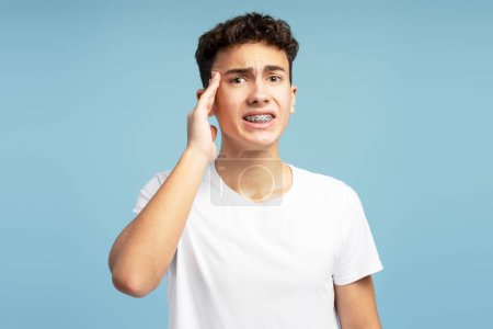 Photo for Sad boy with braces touching head, having migraine, depression, looking at camera. Upset teenager having headache isolated on blue background. Health care concept - Royalty Free Image