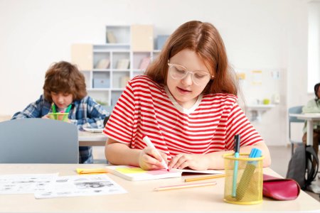 Téléchargez les photos : Adolescente dans des lunettes étudiant en classe et écrivant dans un cahier pendant les cours à l'école. Concept d'apprentissage pour enfants - en image libre de droit