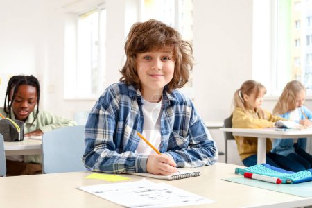 Téléchargez les photos : Écolier souriant assis au bureau et écrivant dans un cahier d'exercices avec un camarade de classe assis en arrière-plan en classe. Concept d'éducation. Retour à l'école - en image libre de droit
