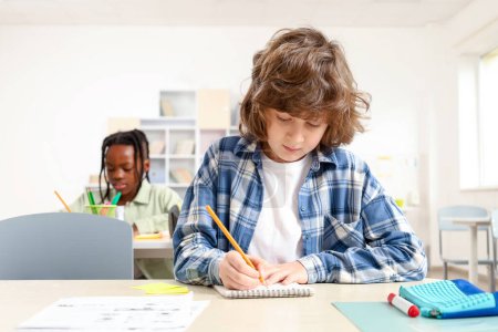 Photo for Schoolboy sitting at classroom and doing exam with his classmates on the background. Education concept. Back to school - Royalty Free Image