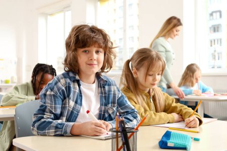 Photo for Happy boy sitting at the desk next to his classmate while teacher helping pupils on the background. Education, learning and people concept - Royalty Free Image