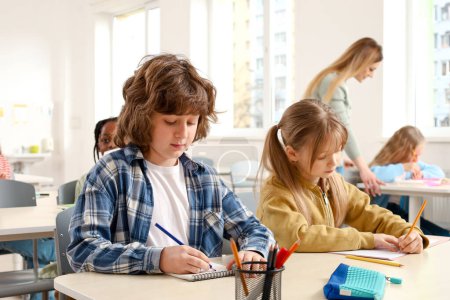 Photo for Schoolboy writing test and sitting at the desk next to girl, teacher helping kids on the background Education, learning and people concept - Royalty Free Image