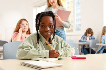Téléchargez les photos : Un écolier afro-américain souriant écrit dans le cahier alors qu'il était assis en classe, enseignant en arrière-plan. Éducation, apprentissage et concept des personnes - en image libre de droit