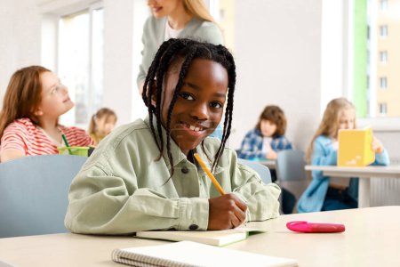 Photo for Smiling African American boy writing in the notebook and sitting at the desk in classroom, classmates on the background. Education, learning and people concept - Royalty Free Image