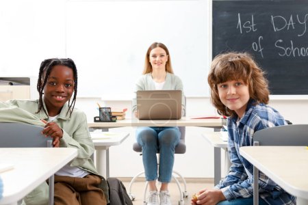 Téléchargez les photos : Heureux élèves assis au bureau dans la salle de classe avec l'enseignant à l'aide d'un ordinateur portable sur le fond. Éducation, apprentissage et concept des personnes - en image libre de droit