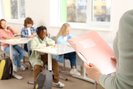 Photo for Cropped photo of teacher with folder during the lesson, pupils sitting at the desk in the classroom. Education, learning and people concept - Royalty Free Image