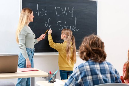 Photo for Schoolgirl writing something on blackboard in classroom while teacher standing next to her. Education concept. Back to school - Royalty Free Image