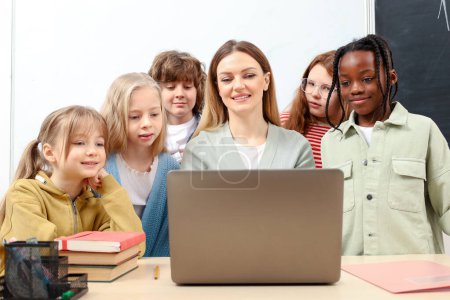 Photo for Pupils standing near the teacher in classroom, using laptop and looking at screen. Education, learning and people concept - Royalty Free Image