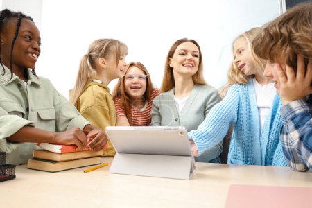 Photo for Smiling pupils standing near the teacher during the lesson, using digital tablet in classroom. Education, learning and people concept - Royalty Free Image