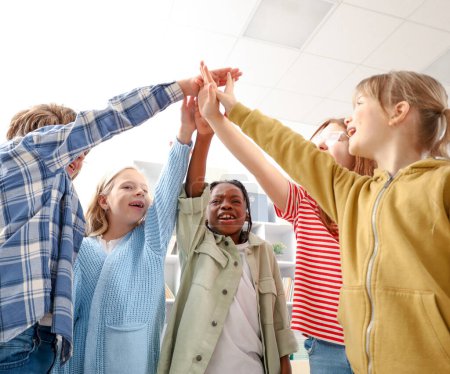 Foto de Retrato de niños felices de la escuela apilándose las manos en el aula, mirando hacia arriba. Vista de ángulo bajo. Unidad, concepto de amistad - Imagen libre de derechos