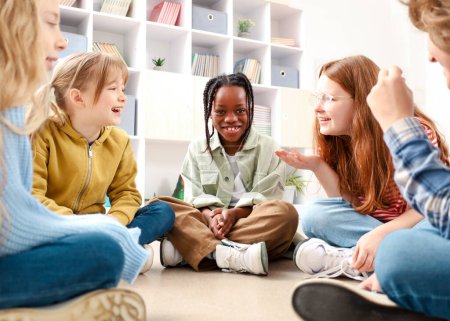 Foto de Niña pelirroja hablando con sus compañeros de clase mientras está sentada en el suelo en el aula de la escuela. Concepto educativo. Regreso a la escuela - Imagen libre de derechos
