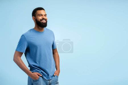 Téléchargez les photos : Heureux jeune homme afro-américain barbu cherchant à copier l'espace tout en posant en studio, isolé sur fond bleu. Concept de style de vie - en image libre de droit