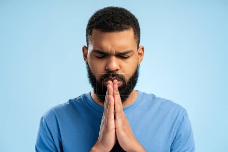 Téléchargez les photos : Portrait d'homme afro-américain barbu en t-shirt priant debout en studio, isolé sur fond bleu. Concept de style de vie - en image libre de droit