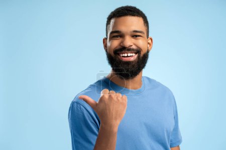 Téléchargez les photos : Heureux homme afro-américain barbu posant et pointant vers le côté avec le pouce vers le haut en studio, isolé sur fond bleu. Concept de style de vie - en image libre de droit