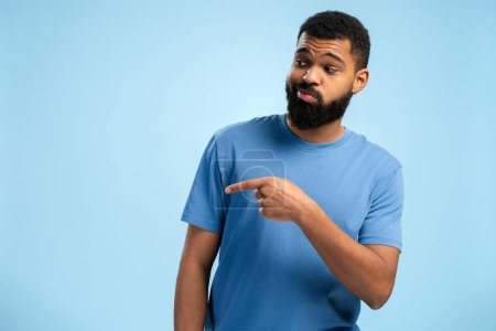 Photo for Bearded African American man posing in t shirt while pointing by finger to the side isolated on blue background. People lifestyle concept - Royalty Free Image