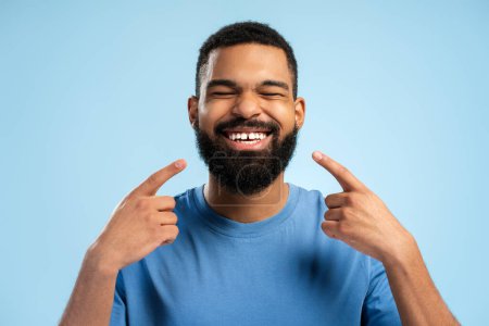 Photo for Smiling African American bearded man posing with closed eyes while man pointing with hands at his teeth, isolated on blue background. People lifestyle concept - Royalty Free Image