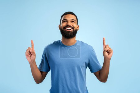 Photo for Smiling young African American man looking up while pointing by fingers to copy space, isolated on blue background. People lifestyle concept - Royalty Free Image