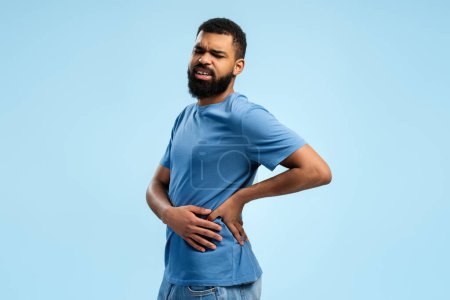 Photo for Sick bearded African American man standing and touching his back, having problems with kidney, isolated on blue background. People lifestyle concept - Royalty Free Image