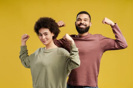 Foto de Feliz hombre y mujer afroamericanos mostrando los músculos de los brazos y mirando a la cámara en el estudio, aislados en el fondo amarillo. Fitness, concepto de deportistas - Imagen libre de derechos