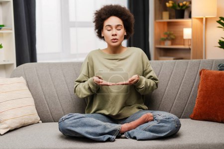 Téléchargez les photos : Jeune femme afro-américaine les yeux fermés pratiquant le yoga méditer à la maison tout en étant assis sur le canapé. Concept de mode de vie sain - en image libre de droit