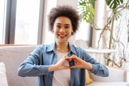 Photo for Smiling beautiful African American young woman showing heart sign looking at camera, gesturing, communicating with sign language while sitting on comfortable couch. Lifestyle concept - Royalty Free Image
