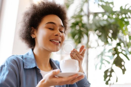 Foto de Feliz sonriente mujer afroamericana con el pelo rizado sosteniendo taza de café, oliendo aroma fragante primer plano de espacio de copia. Concepto de desayuno, publicidad - Imagen libre de derechos