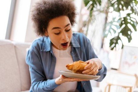 Photo for Excited, attractive, hungry African American woman with curly hair holding tasty croissant, eating while sitting on comfortable sofa in cafe. Concept of morning routine, breakfast - Royalty Free Image