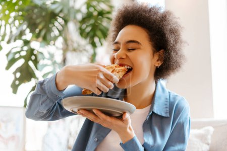 Foto de Hermosa y auténtica joven afroamericana comiendo croissant fresco, con los ojos cerrados, disfrutando mientras está sentada en una acogedora habitación. Concepto de comida, desayuno - Imagen libre de derechos
