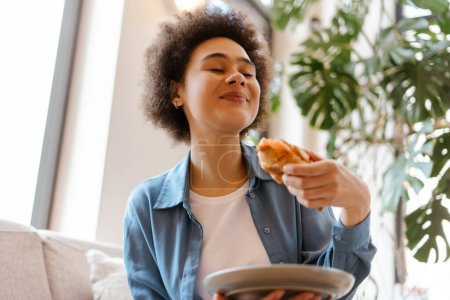 Foto de Sonriente mujer afroamericana alegre con el pelo rizado sosteniendo croissant fresco, comiendo con los ojos cerrados disfrutando sentado en una habitación acogedora. Concepto de comida, desayuno, rutina matutina - Imagen libre de derechos