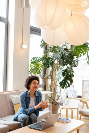 Téléchargez les photos : Belle femme afro-américaine, pigiste, hipster élégant à l'aide d'un ordinateur portable, manger un croissant savoureux tout en étant assis sur un canapé confortable dans un café moderne. Concept de travail à distance, déjeuner, pause - en image libre de droit